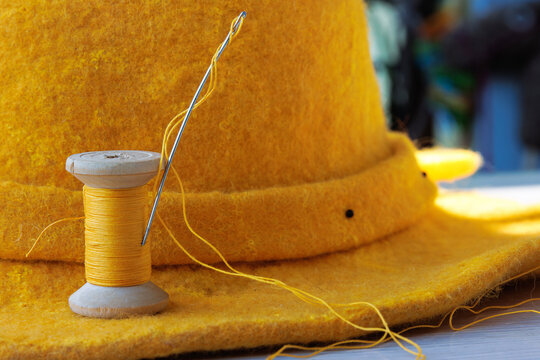 Close up of a yellow thread spool with a sewing needle and loose thread resting on a yellow felt hat brim.