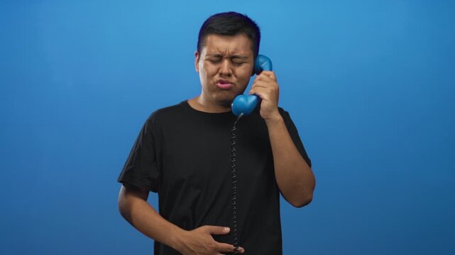 Young man holding blue handset to ear in studio with closed eyes and pained expression; discomfort awkward call.