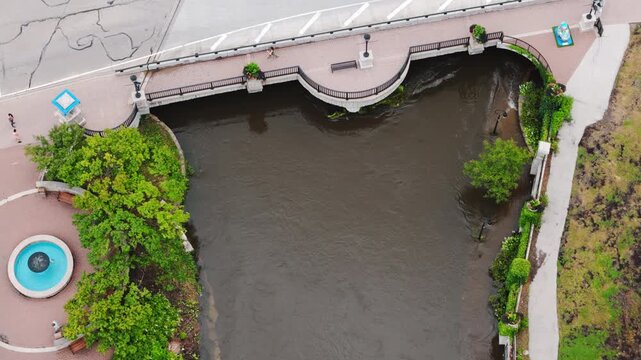 Aerial view of a swollen river flowing through a city after heavy rainfall. High water levels fill the river channel near buildings, bridge, walkways.