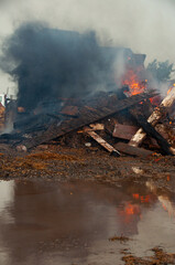 A demolition site scene with charred wood, rubble, and rising smoke. Heavy machinery sits in the background as flames and dust create a hazy, dramatic industrial atmosphere.