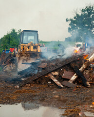 A yellow bulldozer sits empty behind debris burning at a demolition site as flames and smoke rise. Muddy water reflects the machine and fire, capturing industrial energy amid destruction. © Matt