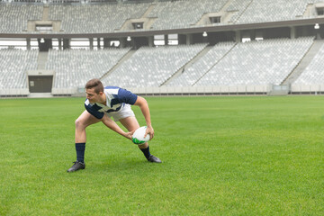 Rugby player crouching low and holding white-and-green ball on grass pitch, empty stands visible