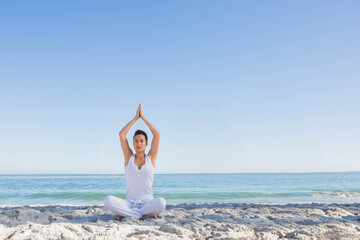 Naklejka premium Woman practicing yoga and meditating while sitting cross legged on sand near ocean in white