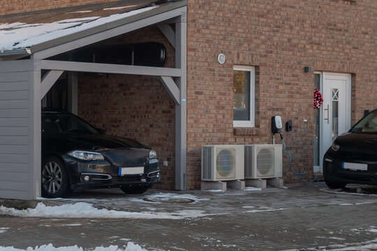 Exterior view of a modern brick residential house featuring a wall-mounted electric vehicle charger and outdoor air source heat pump units.