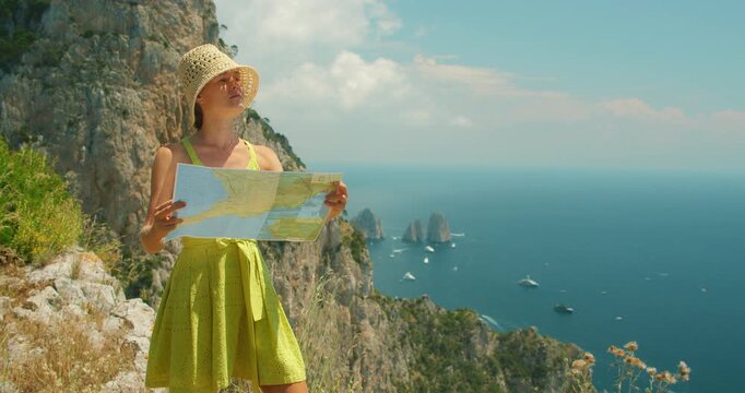 A woman with a sun hat studies a map on a cliff overlooking the Faraglioni sea stacks in Capri Italy