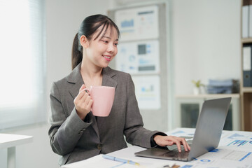 Fototapeta premium Young Asian businesswoman smiling, drinking coffee, and typing on a laptop computer while focusing on her work and financial data documents on a desk in a modern office