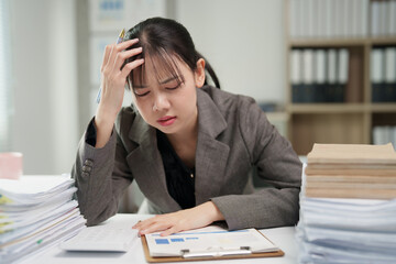 Young asian businesswoman sitting at desk in an office, holding her head experiencing stress, headache, and burnout from excessive workload and pressure, piles of documents surrounding her