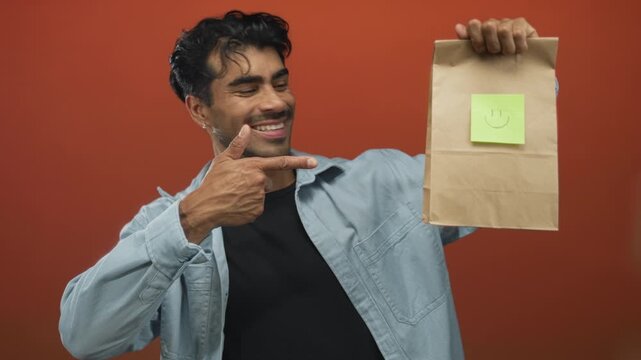 Man holding a brown paper bag with a green sticky note smiley and pointing finger to it against orange studio wall; happiness.