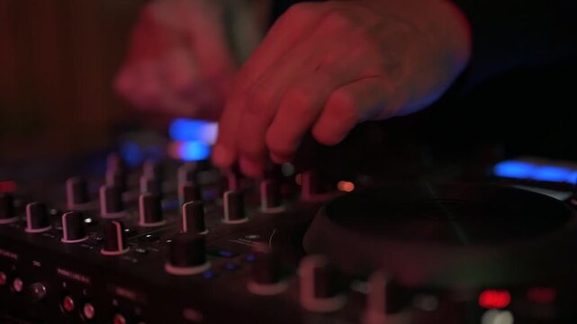 Close up of a professional dj mixing music, adjusting knobs and faders on a soundboard console in a nightclub with vibrant red and blue lights reflecting on the equipment's surface