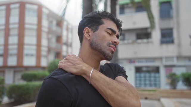 Man touches neck and winces on sunlit city street by building facade wearing a black shirt; discomfort.
