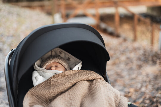 A sleeping newborn baby wrapped in a warm blanket and wearing a knit hat in a stroller outdoors.