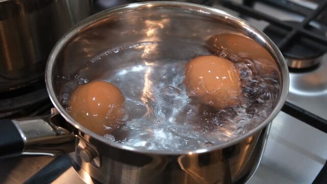 Two eggs boiling in a stainless steel pot of bubbling water on a kitchen stovetop, showing active cooking and heat from the burner. Home cooking, boiling process, and food preparation routine.