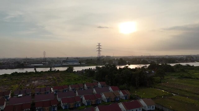 A peaceful evening view with rows of houses along the river, distant industrial landscapes, and tall electricity pylons on the horizon. 
