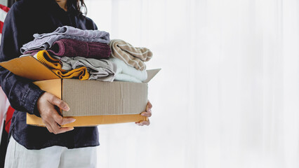 Female hands holding a cardboard box filled with used clothing. Concept of clothes donation, charity, sustainability, recycling, and home decluttering.