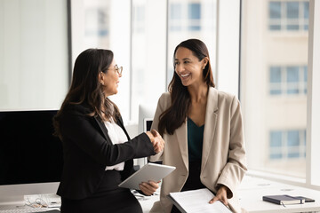 Two happy diverse professional women colleagues shaking hands, finalizing meeting in modern coworking office, expressing recognition and respect for work done. Successful collaboration, companionship © fizkes
