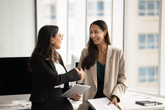 Two happy diverse professional women colleagues shaking hands, finalizing meeting in modern coworking office, expressing recognition and respect for work done. Successful collaboration, companionship