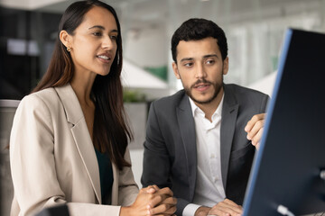 Two colleagues reviewing platform, discussing joint project details, strategizing seated at table looking pc monitor with focused expression. Mentorship, client and manager formal meeting, teamwork