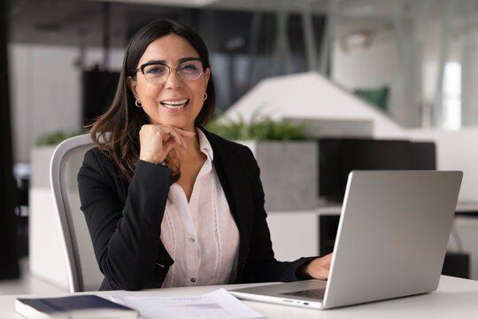 Portrait of mature 60s businesslady boss, female staff member in glasses, elegant business attire sits at desk using laptop, posing for camera with contented smile, exude professionalism and authority