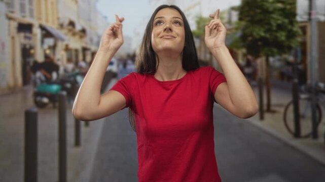 Woman crossing fingers with hands raised on street, eyes closed and smiling, wearing red t shirt; hopeful luck.