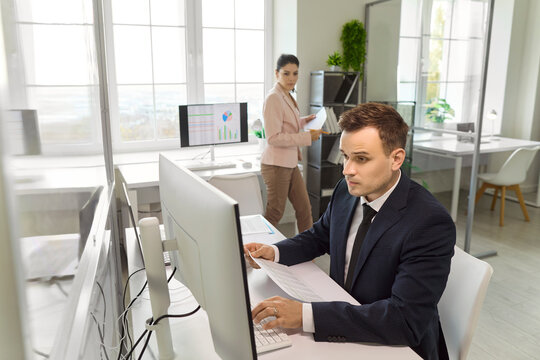 Businessman at computer conducts analysis of data. He types and checks a printed report while charts glow on the monitor in a bright workspace. Concept: focused productivity and accuracy.
