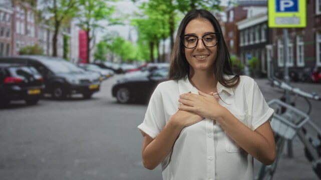 Young hispanic woman hands on chest smiling on a street with parked cars and a bicycle rack; gratitude connection.
