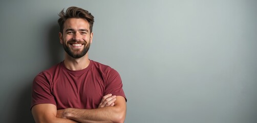 Bearded man with crossed arms smiles warmly. He wears a red tee shirt, standing casually against a plain grey wall. Friendly, approachable, confident pose.