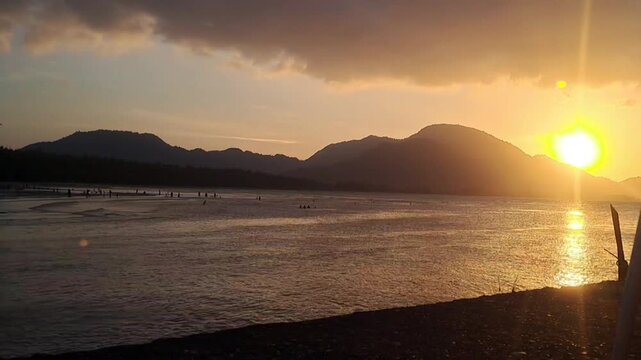 Time lapse of a seaside sunset with moving clouds, calm water, boats, and mountains in the background during golden hour.