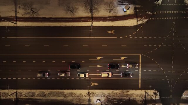 Aerial top-down view of vehicles waiting at a traffic light on a multi-lane road in Vilnius at night during winter, with snow-covered sidewalks and clear lane markings.