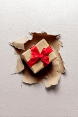 a gift box with a red ribbon wrapped around it, peeking out from the hole in a ripped brown paper background.