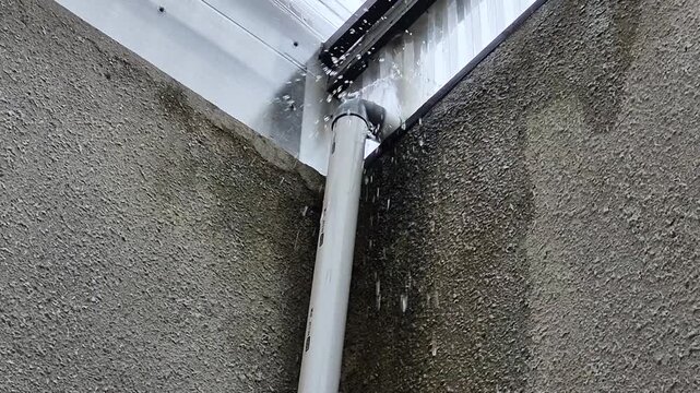 Leaking rain gutter pipe during heavy rainfall, showing drainage problem, water damage risk, and home maintenance issue