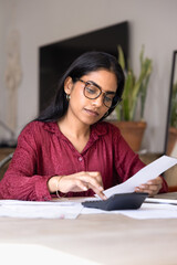 Focused young Indian woman in stylish glasses calculating regular payments, budget, checking...