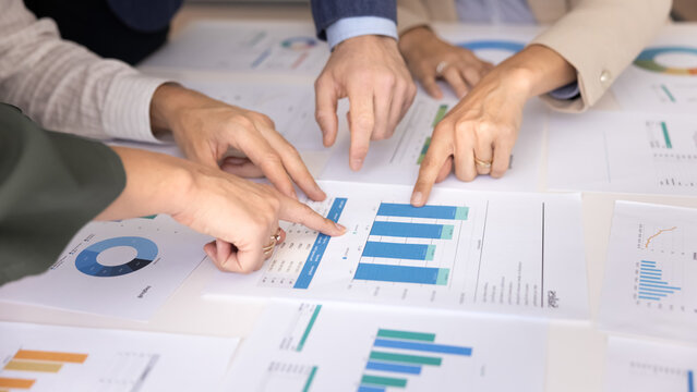 Multiple hands pointing at various printed bar, pie charts and graphs laid out on table, close up view. Unknown teammates discussing business or financial data shown on infographic papers documents