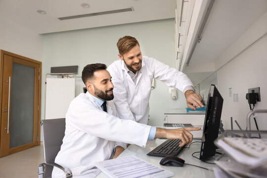 Digital healthcare achievement. Two young male doctors mentor and intern point at desktop monitor with smiles make diagnosis together review correct treatment result confirm positive patient progress