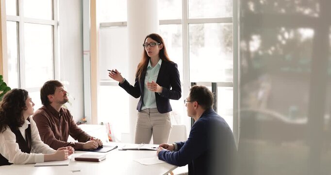 Adult 30s Caucasian businesswoman standing and explaining project details, presenting strategy and ideas to group of corporate members during team briefing or business meeting in modern office