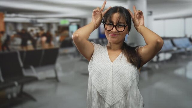 Brunette woman smiling with both hands making peace signs by her head wearing white top in busy airport terminal, glasses and lip piercing visible; playful travel moment.
