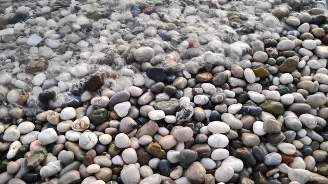 Close up view of sea waves washing over colorful rounded pebbles on beach. White foam and surf flowing between stones. Mediterranean beach texture footage