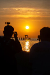 Fototapeta premium Silhouetted photographers capturing golden sunset over pier and calm sea at Bang Pu in Samut Prakan, Thailand. Popular spot to relax, enjoy sunrise and sunsets.