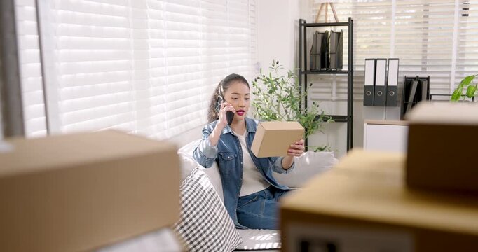 Asian woman small business owner sits on sofa holding smartphone, discusses customer order details, reviews online shopping parcel box status, coordinates e-commerce order delivery planning work call