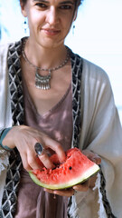 A beautiful brunette in a bohemian style relaxes outdoors in the summer, holding a large slice of watermelon against a green forest backdrop.