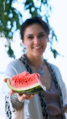A beautiful brunette in a bohemian style relaxes outdoors in the summer, holding a large slice of watermelon against a green forest backdrop.