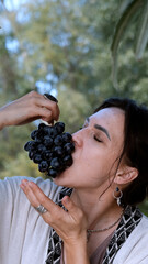 A beautiful brunette in a bohemian style relaxes outdoors in summer against a backdrop of a green forest, holding a large bunch of juicy black grapes