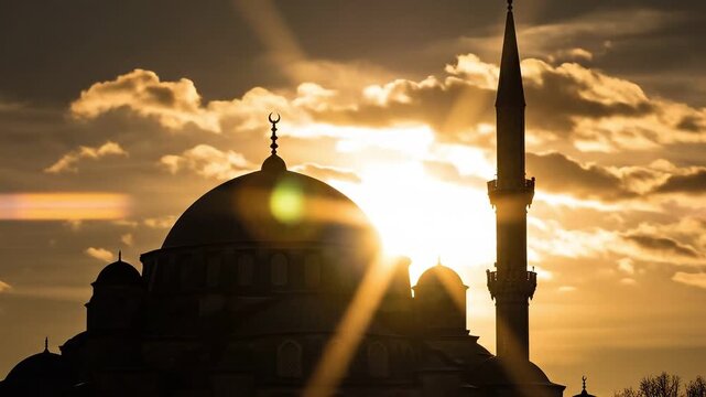 Silhouette of a grand mosque with a tall minaret against a bright golden sunset sky featuring dramatic moving clouds and warm light