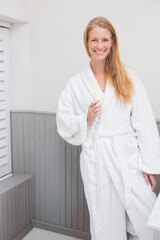 Mid-adult woman standing in small bathroom wearing white terry bathrobe holding toothbrush