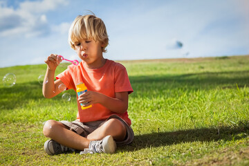 Fototapeta premium Boy sitting cross-legged on hill blowing bubbles with pink wand, yellow bottle, wearing orange tee