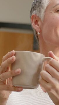 Vertical video: Lifting mug to mouth, mature woman in white top sipping drink at home, showing ring