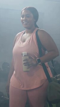 Vertical video: Walking lady in pink entering gym from left to lift weights, holding bag and bottle
