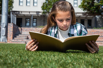 Girl reading large book, lying on grassy lawn outside brick building under sunny sky