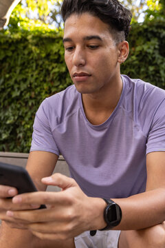 Man in early 20s sitting on garden bench wearing sportswear, checking smartphone, fitness watch