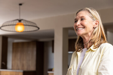 Senior woman standing in kitchen with warm pendant lamp over wood cabinets, copy space © wavebreak3