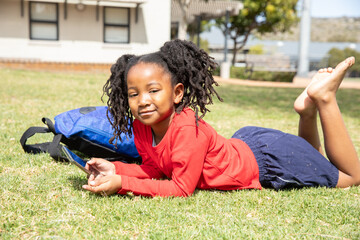 African american girl child lying on grassy lawn in front of school using smartphone and backpack